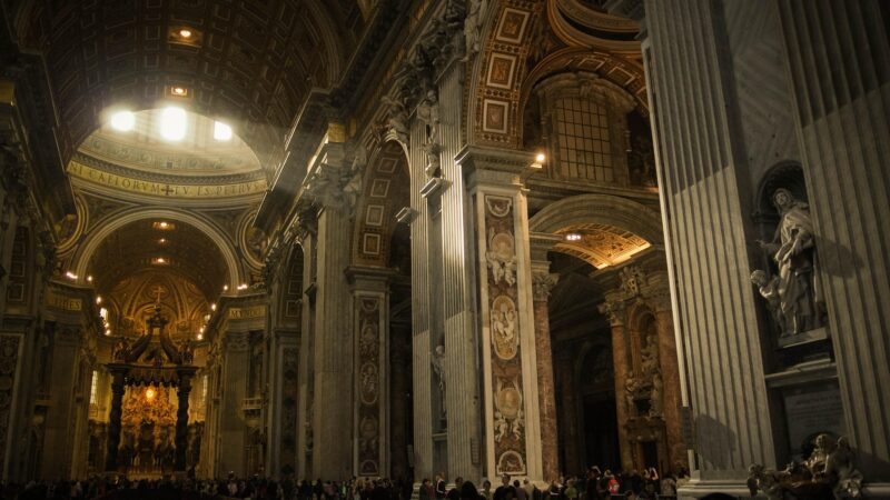 Interior da Basílica de São Pedro no Vaticano mostrando nave central com colunas monumentais, teto decorado dourado, baldaquino de Bernini e turistas visitando.