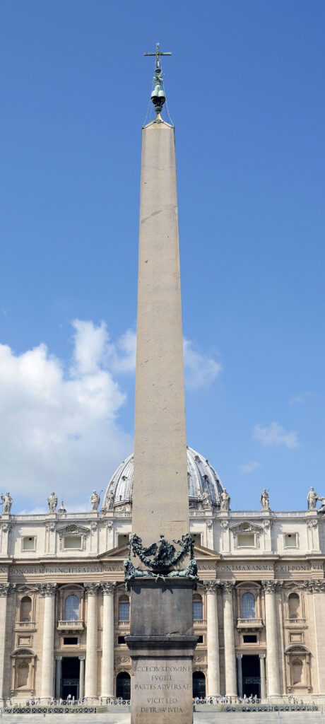 Obelisco do Vaticano na Praça de São Pedro com a cruz no topo e a fachada com a cúpula da Basílica de São Pedro ao fundo, céu azul.