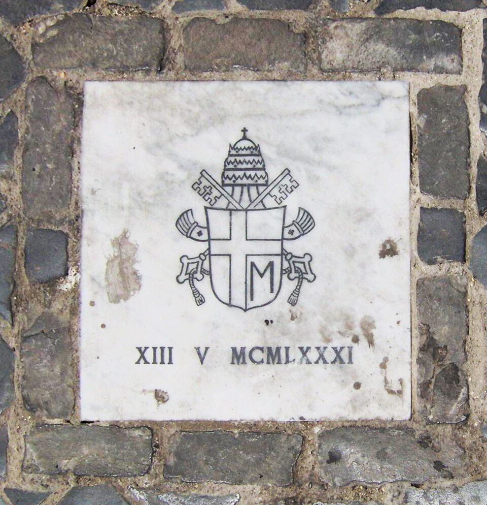 Placa comemorativa do atentado ao Papa João Paulo II em 13 de maio de 1981 na Praça de São Pedro, com brasão papal e inscrição "XIII V MCMLXXXI"