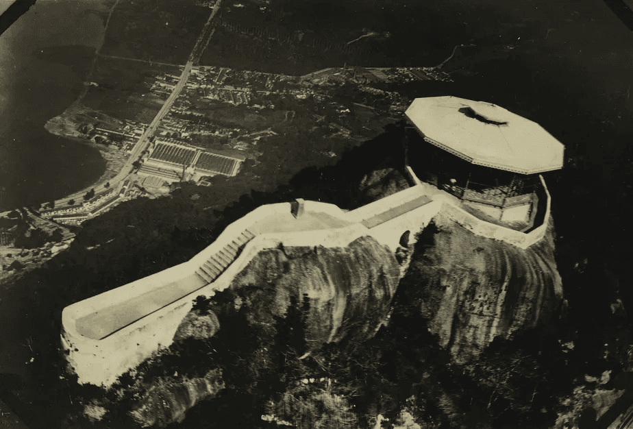 Fotografia aérea histórica em preto e branco do Morro da Urca no Rio de Janeiro, mostrando a estação octogonal original do Bondinho do Pão de Açúcar e a encosta rochosa, início do século XX