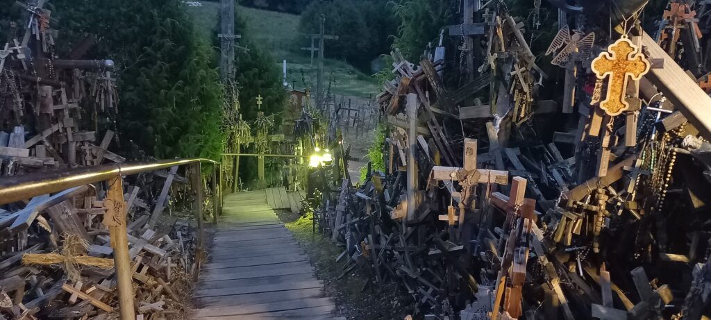 Caminho de madeira iluminado atravessando a Colina das Cruzes em Šiauliai, Lituânia, com milhares de cruzes de madeira próximas e rosários pendurados