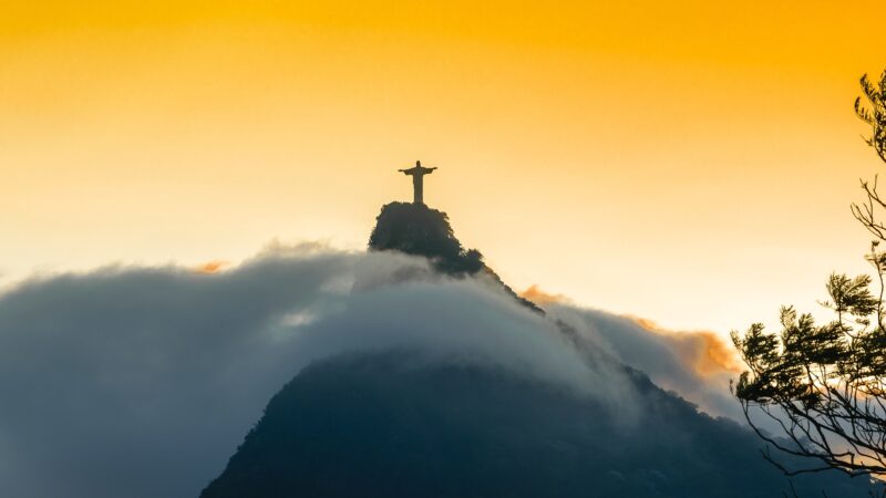 Silhueta do Cristo Redentor no topo do Corcovado ao pôr do sol, com nuvens envolvendo a montanha e céu dourado, Rio de Janeiro Brasil