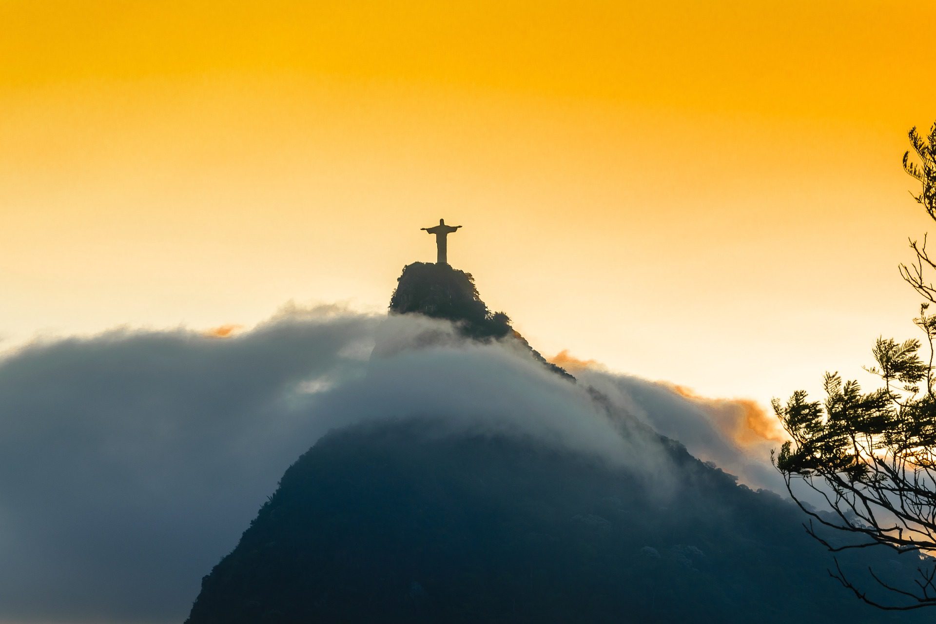 Silhueta do Cristo Redentor no topo do Corcovado ao pôr do sol, com nuvens envolvendo a montanha e céu dourado, Rio de Janeiro Brasil