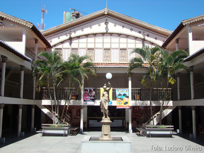 Pátio interno de edifício histórico brasileiro com arquitetura colonial, estátua central, palmeiras e galerias com colunas, céu azul
