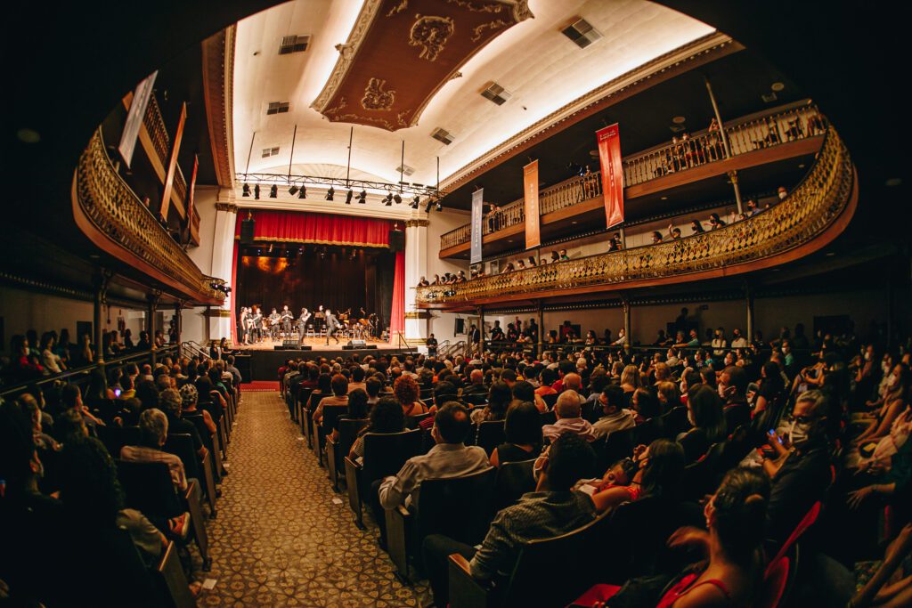 Espetáculo musical no Theatro Alberto Maranhão em Natal RN com plateia lotada, palco com cortina vermelha, balcões dourados ornamentados e teto decorado