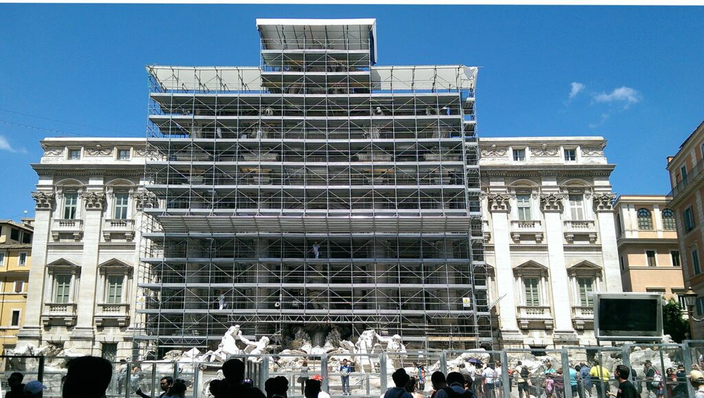 Fontana di Trevi em Roma coberta por andaimes durante restauração, com turistas observando a fonte barroca em obras sob céu azul