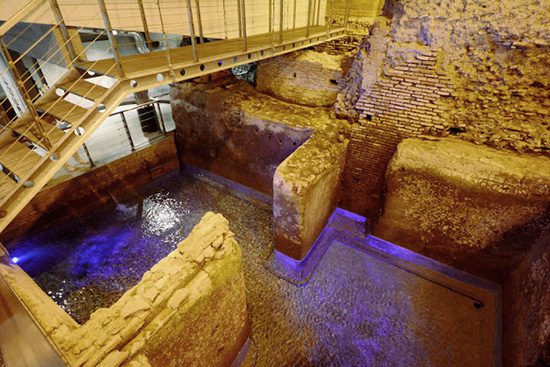 Vicus Caprarius em Roma mostrando estruturas antigas do aqueduto Aqua Virgo com água, paredes de tijolos romanos, passarela moderna e iluminação azul subterrânea