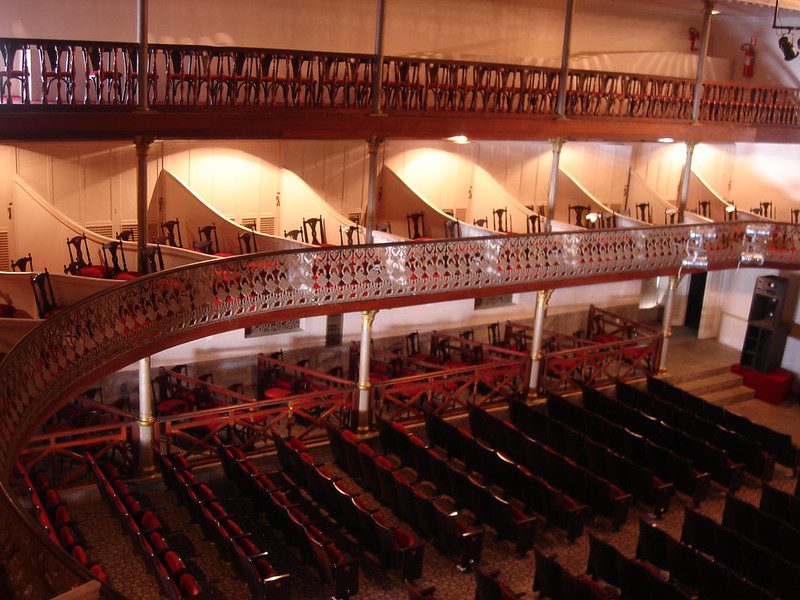 Interior do Theatro Alberto Maranhão em Natal RN mostrando plateia com cadeiras vermelhas, balcões ornamentados com grades de ferro trabalhado e arquitetura eclética do teatro histórico
