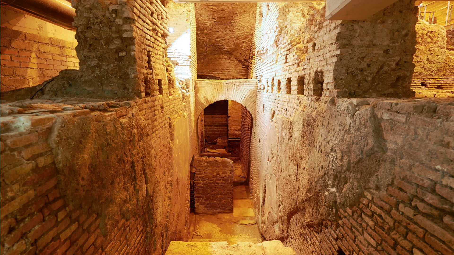 Vicus Caprarius - Ruínas arqueológicas subterrâneas sob a Fontana di Trevi em Roma, mostrando paredes de tijolos romanos antigos, corredor estreito e arco com iluminação dourada