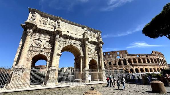 Arco de Constantino em Roma com Coliseu ao fundo mostrando arco do triunfo romano antigo, relevos históricos, turistas visitando e pinheiro italiano sob céu azul