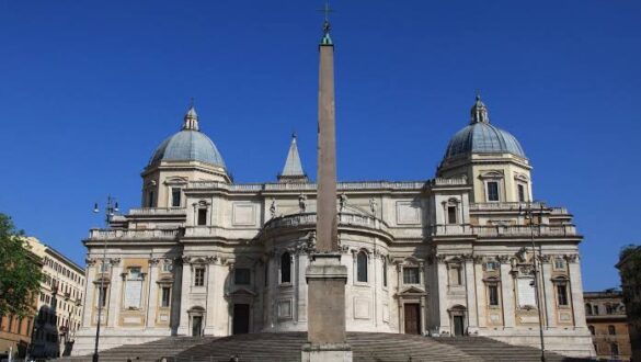 Basílica de Santa Maria Maggiore em Roma com duas cúpulas simétricas azuis, obelisco egípcio alto no centro da praça, escadaria monumental e fachada barroca sob céu azul intenso