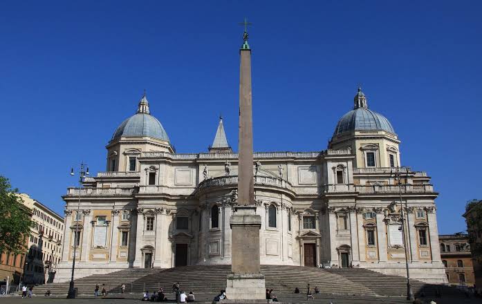 Basílica de Santa Maria Maggiore em Roma com duas cúpulas simétricas azuis, obelisco egípcio alto no centro da praça, escadaria monumental e fachada barroca sob céu azul intenso