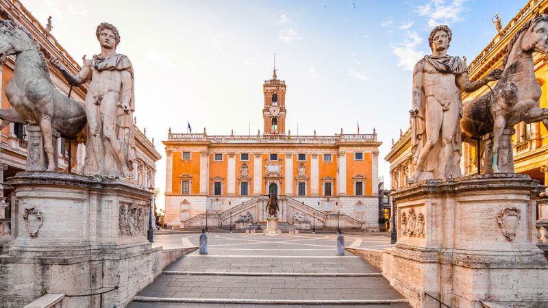 Piazza del Campidoglio em Roma mostrando estátuas dos Dioscuri (Castor e Pólux) com cavalos, Palazzo Senatorio com fachada laranja e torre do relógio, escadaria monumental e praça simétrica projetada por Michelangelo