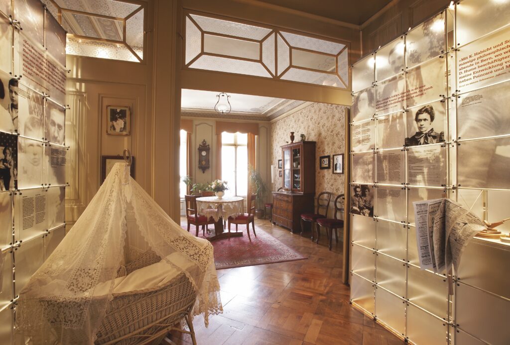 Interior da Einstein Haus em Berna Suíça mostrando berço de vime com dossel de renda, painéis expositivos com fotografias históricas de Albert Einstein e família, e sala mobiliada com móveis de época do início do século XX