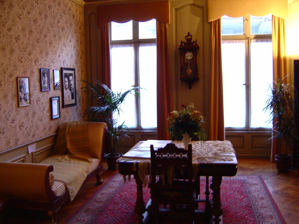 Interior do apartamento de Albert Einstein na Einstein Haus Berna mostrando sala mobiliada com móveis de época, sofá estofado, mesa de madeira entalhada, relógio de parede, fotografias históricas, papel de parede floral e janelas com cortinas