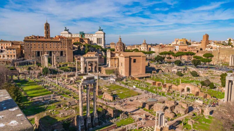 Vista panorâmica do Fórum Romano em Roma Itália mostrando ruínas antigas com colunas, templos, Arco de Septímio Severo, áreas verdes e ao fundo o monumento Vittoriano e skyline da cidade eterna sob céu azul