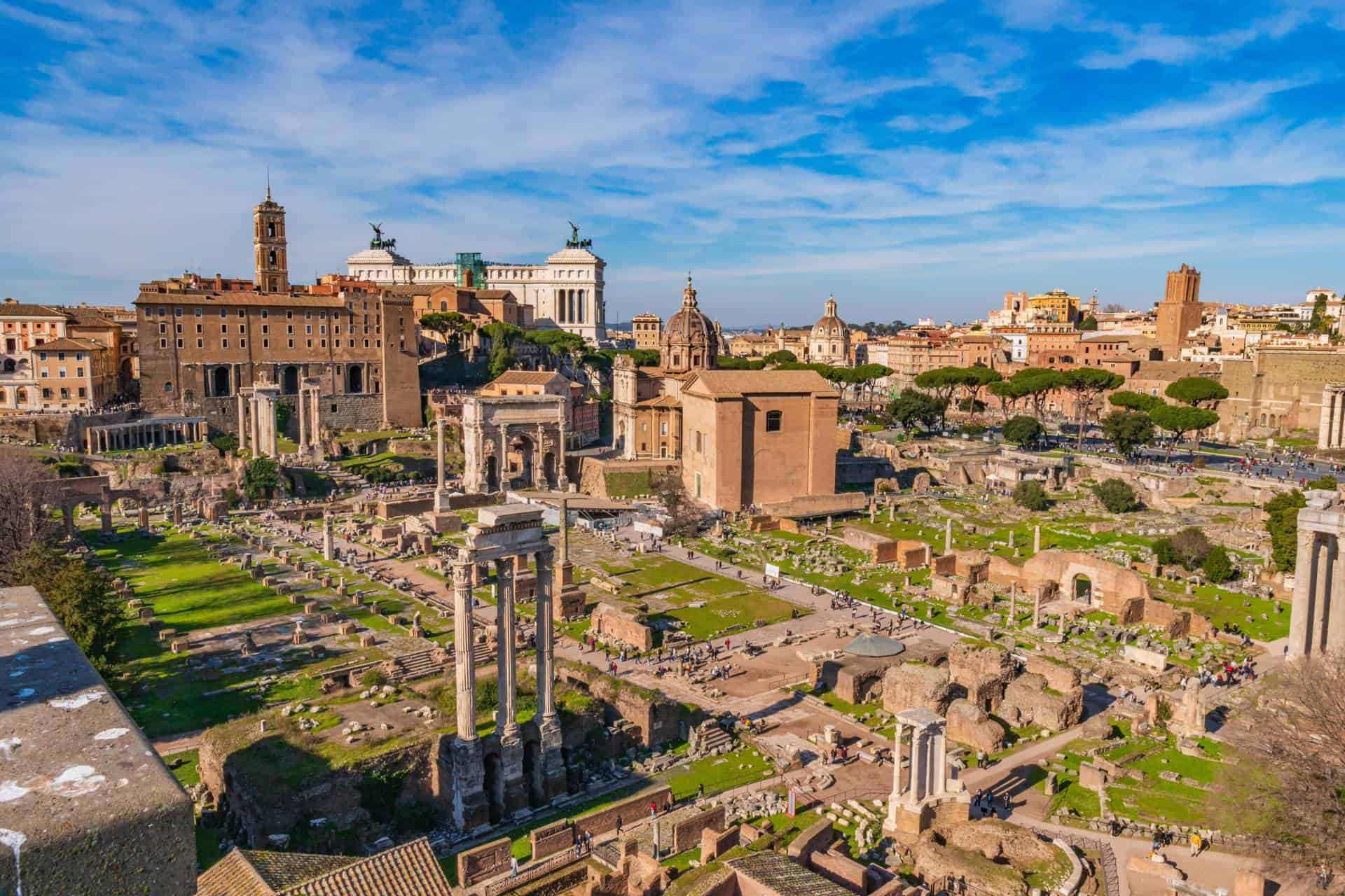 Vista panorâmica do Fórum Romano em Roma Itália mostrando ruínas antigas com colunas, templos, Arco de Septímio Severo, áreas verdes e ao fundo o monumento Vittoriano e skyline da cidade eterna sob céu azul