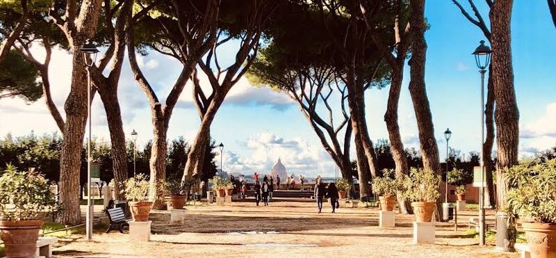 Avenida arborizada com pinheiros italianos em Roma mostrando caminho com vasos de terracota, postes de luz e vista panorâmica para a cúpula da Basílica de São Pedro no Vaticano ao fundo