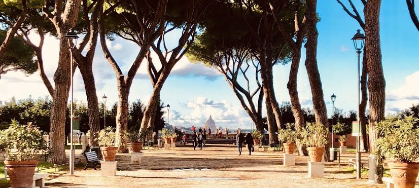 Avenida arborizada com pinheiros italianos em Roma mostrando caminho com vasos de terracota, postes de luz e vista panorâmica para a cúpula da Basílica de São Pedro no Vaticano ao fundo
