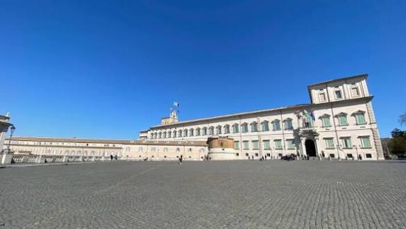 Grande palácio histórico com arquitetura neoclássica, fachada simétrica com múltiplas janelas, torre central, praça ampla de paralelepípedos e céu azul
