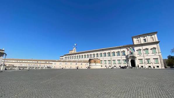 Grande palácio histórico com arquitetura neoclássica, fachada simétrica com múltiplas janelas, torre central, praça ampla de paralelepípedos e céu azul