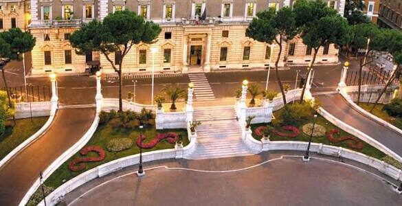 Palácio do Quirinal em Roma à noite mostrando fachada neoclássica iluminada, escadaria monumental central com balaustrada branca, jardins formais com canteiros florais em formato de S, pinheiros italianos e caminhos curvos
