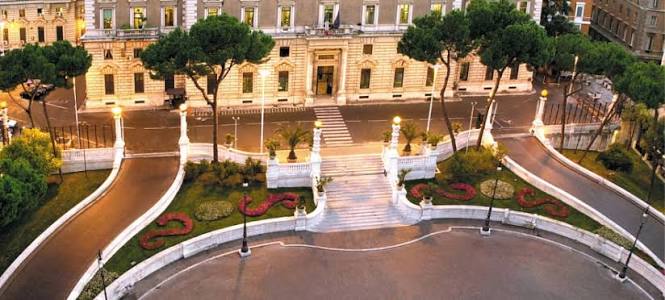 Palácio do Quirinal em Roma à noite mostrando fachada neoclássica iluminada, escadaria monumental central com balaustrada branca, jardins formais com canteiros florais em formato de S, pinheiros italianos e caminhos curvos