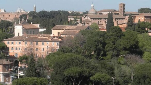 Vista panorâmica de Roma mostrando telhados históricos, edifícios com fachadas alaranjadas, pinheiros-mansos italianos, torre sineira de tijolos e ruínas antigas ao fundo