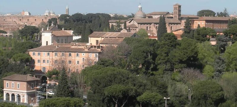 Vista panorâmica de Roma mostrando telhados históricos, edifícios com fachadas alaranjadas, pinheiros-mansos italianos, torre sineira de tijolos e ruínas antigas ao fundo