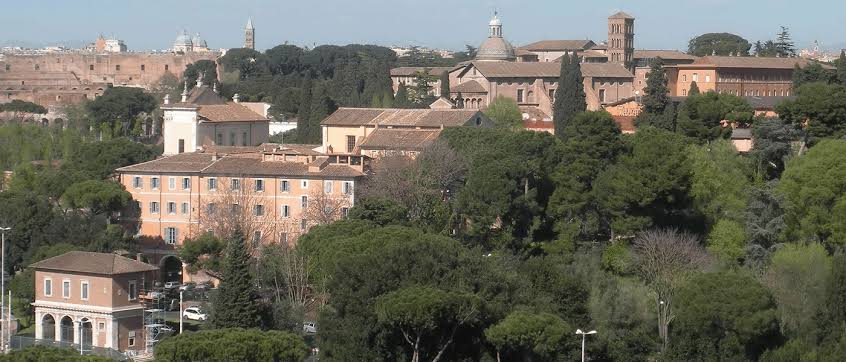 Vista panorâmica de Roma mostrando telhados históricos, edifícios com fachadas alaranjadas, pinheiros-mansos italianos, torre sineira de tijolos e ruínas antigas ao fundo