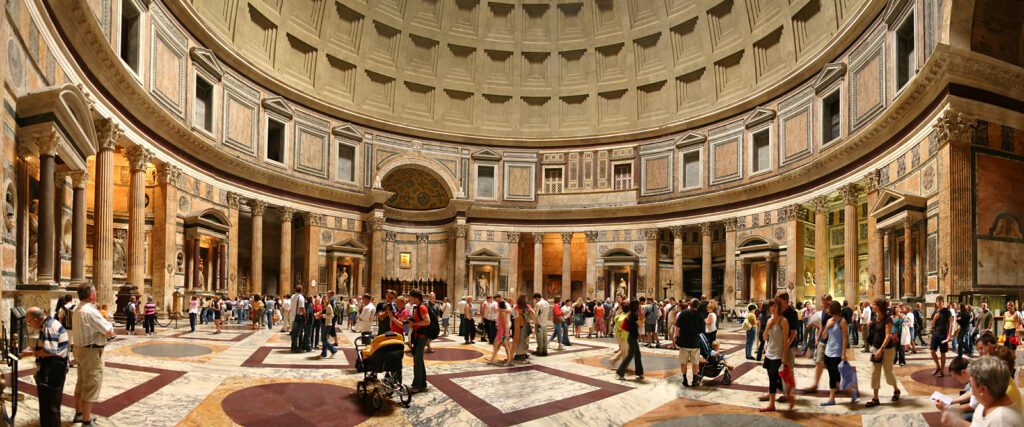 Interior panorâmico do Panteão de Roma mostrando rotunda circular com cúpula de caixotões, paredes decoradas com mármores coloridos, colunas, nichos, piso geométrico e multidão de turistas visitando o monumento