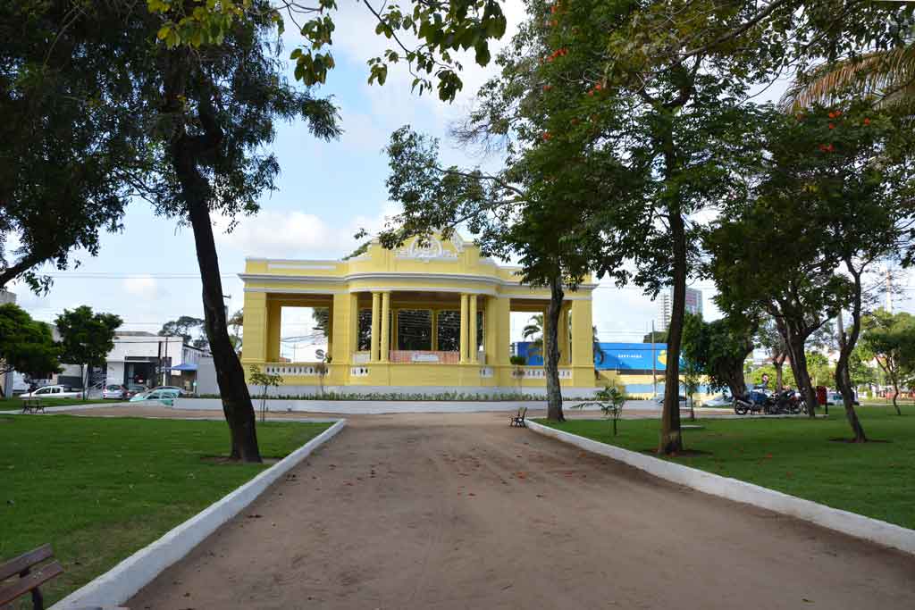 Coreto amarelo com colunas brancas em praça pública brasileira, mostrando arquitetura neoclássica, gramado verde, árvores, caminho de terra e bancos de madeira em área de lazer urbana