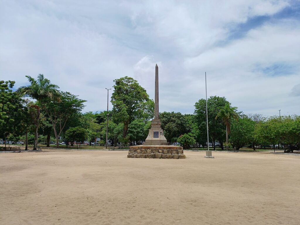 Obelisco monumento em praça pública com base de pedra circular, árvores tropicais e palmeiras ao redor, céu nublado, área de lazer urbana arborizada