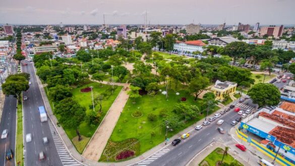 Vista aérea de parque urbano com grande área verde, árvores, jardins, avenidas laterais com tráfego e edifícios ao fundo em cidade brasileira, mostrando planejamento urbano e área de lazer central