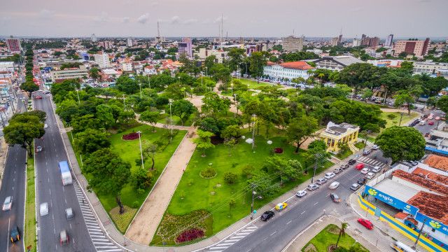Vista aérea de parque urbano com grande área verde, árvores, jardins, avenidas laterais com tráfego e edifícios ao fundo em cidade brasileira, mostrando planejamento urbano e área de lazer central