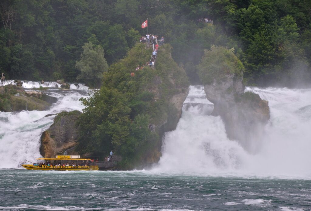 Barco turístico amarelo Felsenfahrt navegando próximo à rocha central das Cataratas do Reno na Suíça, com bandeira suíça hasteada no topo da rocha coberta de vegetação, turistas subindo escadaria e cachoeiras poderosas ao redor com névoa