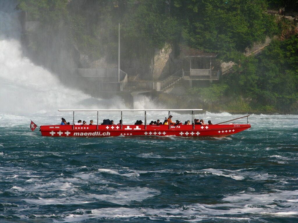 Barco turístico vermelho maendli.ch navegando nas águas turbulentas das Cataratas do Reno na Suíça, com forte névoa da cachoeira, plataforma de observação de pedra e floresta ao fundo