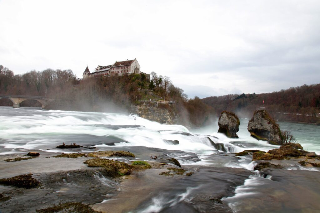 Cataratas do Reno na Suíça com efeito de longa exposição mostrando águas sedosas, Castelo de Laufen no penhasco, rochas centrais cobertas de musgo e ponte de pedra ao fundo em dia nublado de inverno