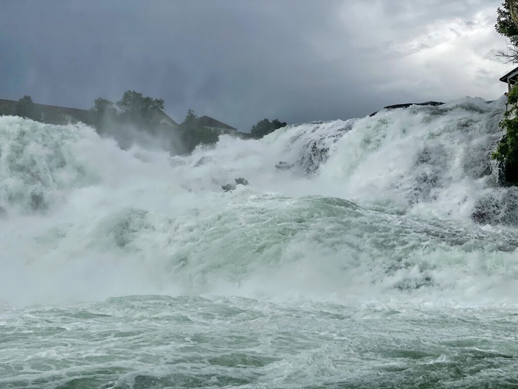 Cataratas do Reno Suíça vista próxima mostrando força bruta da água caindo com intensidade, espuma branca abundante, névoa densa, céu nublado dramático e telhados de edifícios ao fundo