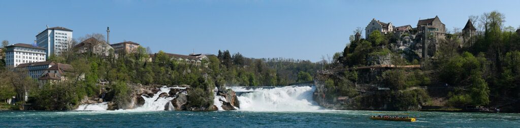 Vista panorâmica das Cataratas do Reno na Suíça mostrando cachoeira em toda extensão, Castelo de Laufen medieval no topo da colina à direita, hotéis e edifícios à esquerda, barco turístico amarelo navegando nas águas turquesa sob céu azul