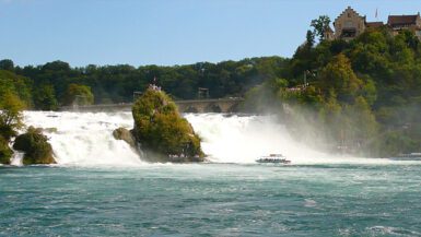 Cataratas do Reno na Suíça mostrando cachoeira poderosa com rochas cobertas de vegetação, barco turístico navegando nas águas turquesa, castelo medieval Schloss Laufen no topo da colina arborizada e névoa das quedas d'água