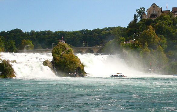 Cataratas do Reno na Suíça mostrando cachoeira poderosa com rochas cobertas de vegetação, barco turístico navegando nas águas turquesa, castelo medieval Schloss Laufen no topo da colina arborizada e névoa das quedas d'água