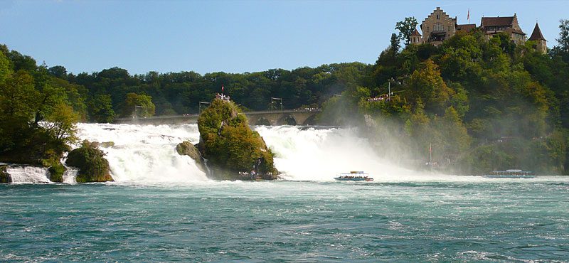 Cataratas do Reno na Suíça mostrando cachoeira poderosa com rochas cobertas de vegetação, barco turístico navegando nas águas turquesa, castelo medieval Schloss Laufen no topo da colina arborizada e névoa das quedas d'água
