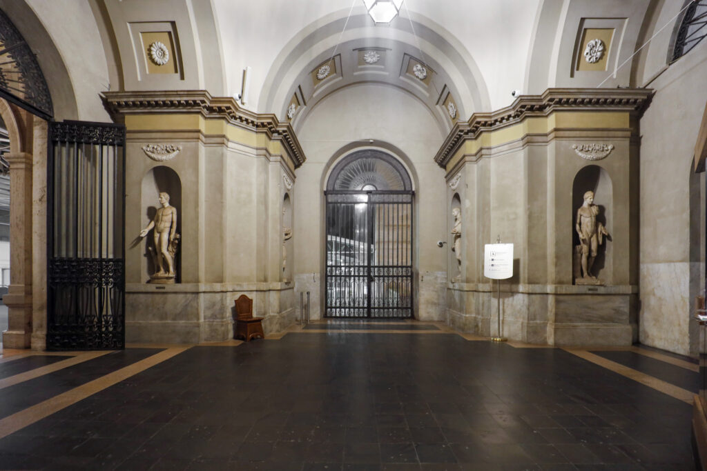 Corredor interno dos Museus do Vaticano com estátuas clássicas em nichos e portões de ferro forjado