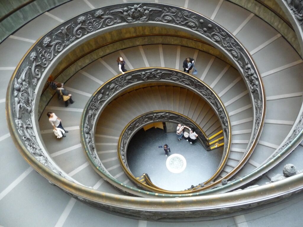 Vista aérea da Escadaria em Espiral de Giuseppe Momo na saída dos Museus do Vaticano, com corrimão decorado em bronze e turistas descendo