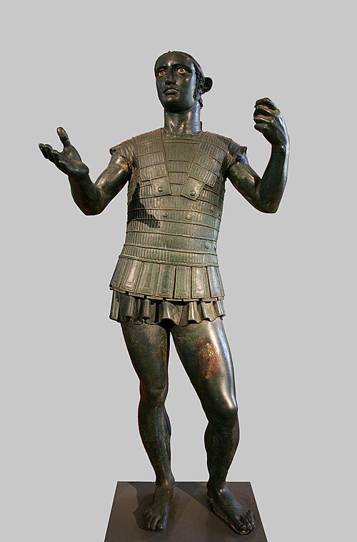 Estátua de bronze etrusca ou romana retratando figura em armadura militar com braços erguidos, acervo dos Museus do Vaticano