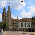 Grossmünster em Zurique Suíça com duas torres românicas, estátua equestre de Hans Waldmann em primeiro plano, ponte de pedestres com turistas, edifícios históricos e céu azul com nuvens