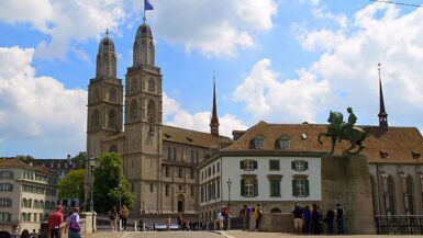 Grossmünster em Zurique Suíça com duas torres românicas, estátua equestre de Hans Waldmann em primeiro plano, ponte de pedestres com turistas, edifícios históricos e céu azul com nuvens