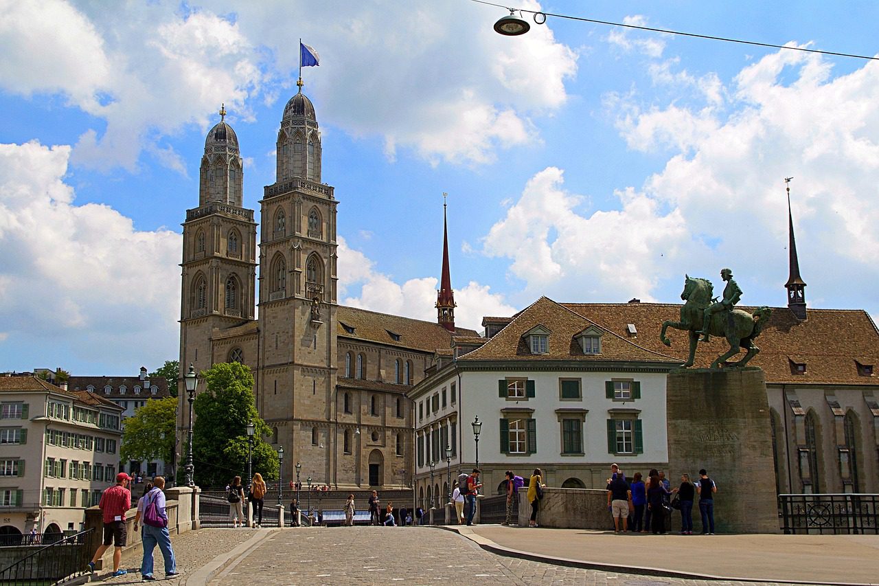 Grossmünster em Zurique Suíça com duas torres românicas, estátua equestre de Hans Waldmann em primeiro plano, ponte de pedestres com turistas, edifícios históricos e céu azul com nuvens