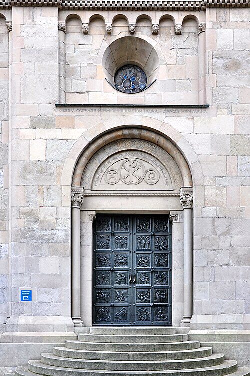 Porta lateral da Grossmünster em Zurique Suíça mostrando portal românico com tímpano decorado com símbolo Chi-Rho, portas de bronze com relevos narrativos, óculo com vitral e inscrição em latim mencionando Zwingli